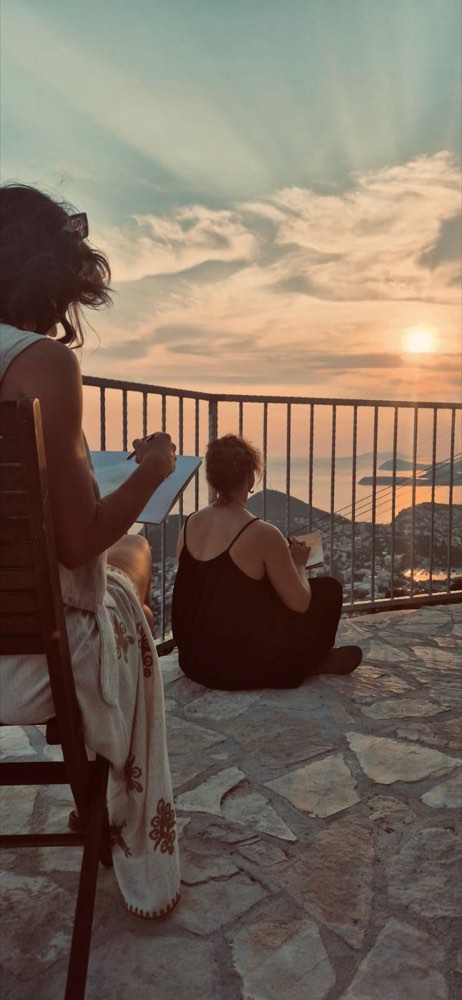 Two people sketching at the railing as the sun sets over the islands.