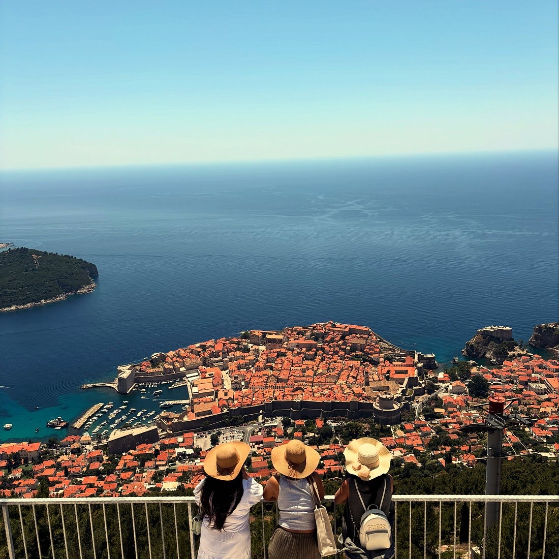 Visitors at the railing of the viewpoint, looking down over Dubrovnik's Old Town and the Adriatic.