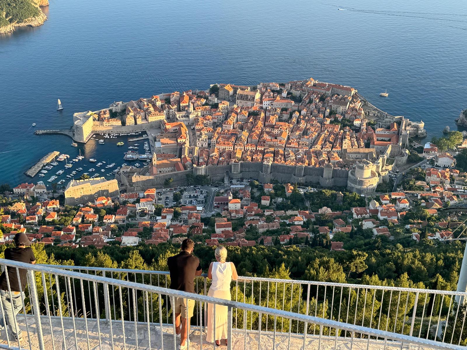 The view from the terrace railing: the walled Old Town of Dubrovnik and its harbour below, Lokrum and the open sea beyond, at golden hour.